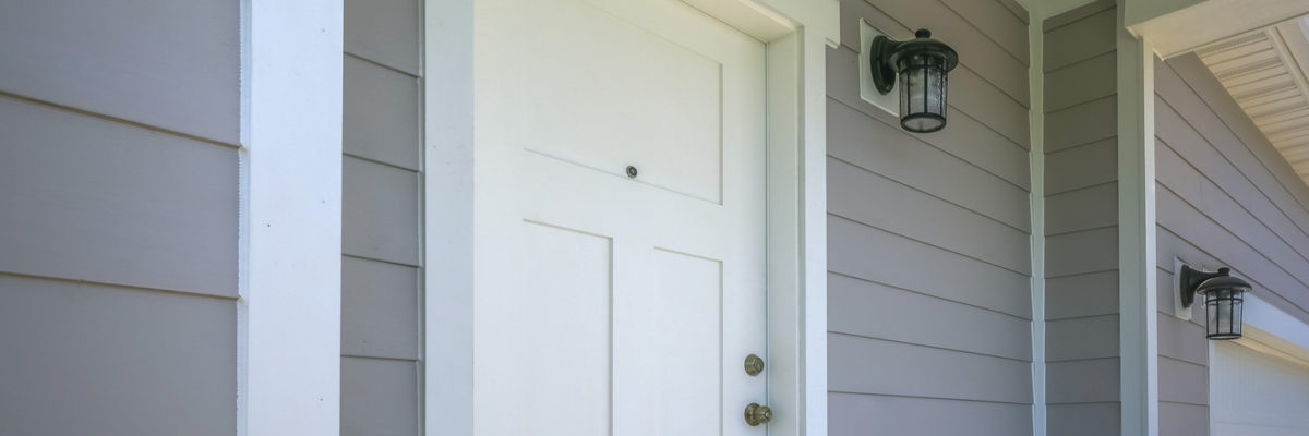 Front door of a home with a lamp on the gray wall. View of a home with a wooden front door and white garage door. Outdoor lamps are mounted on the gray wall with horizontal siding.